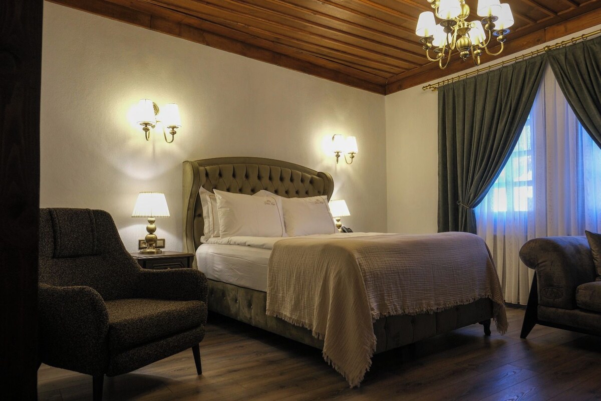 A hotel room with a large bed, two bedside lamps, a chandelier, and a view of the outdoors through curtains.