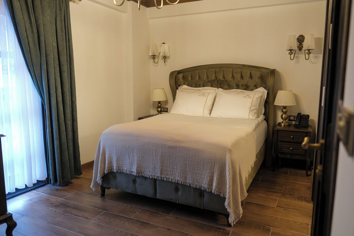 Una cama bien hecha con cabecera capitonada, dos mesitas de noche con lámparas y vista al suelo de madera de la habitación.