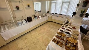 En buffé med olika sorters mat och serveringsredskap uppdukad på ett långt bord, omgivet av hyllor med blommor och ett fönster med gardiner.