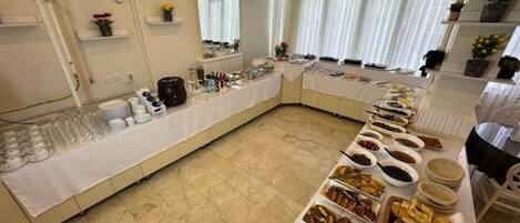 A buffet setup with various food items and utensils on a long table, surrounded by shelves with flowers and a window with curtains.