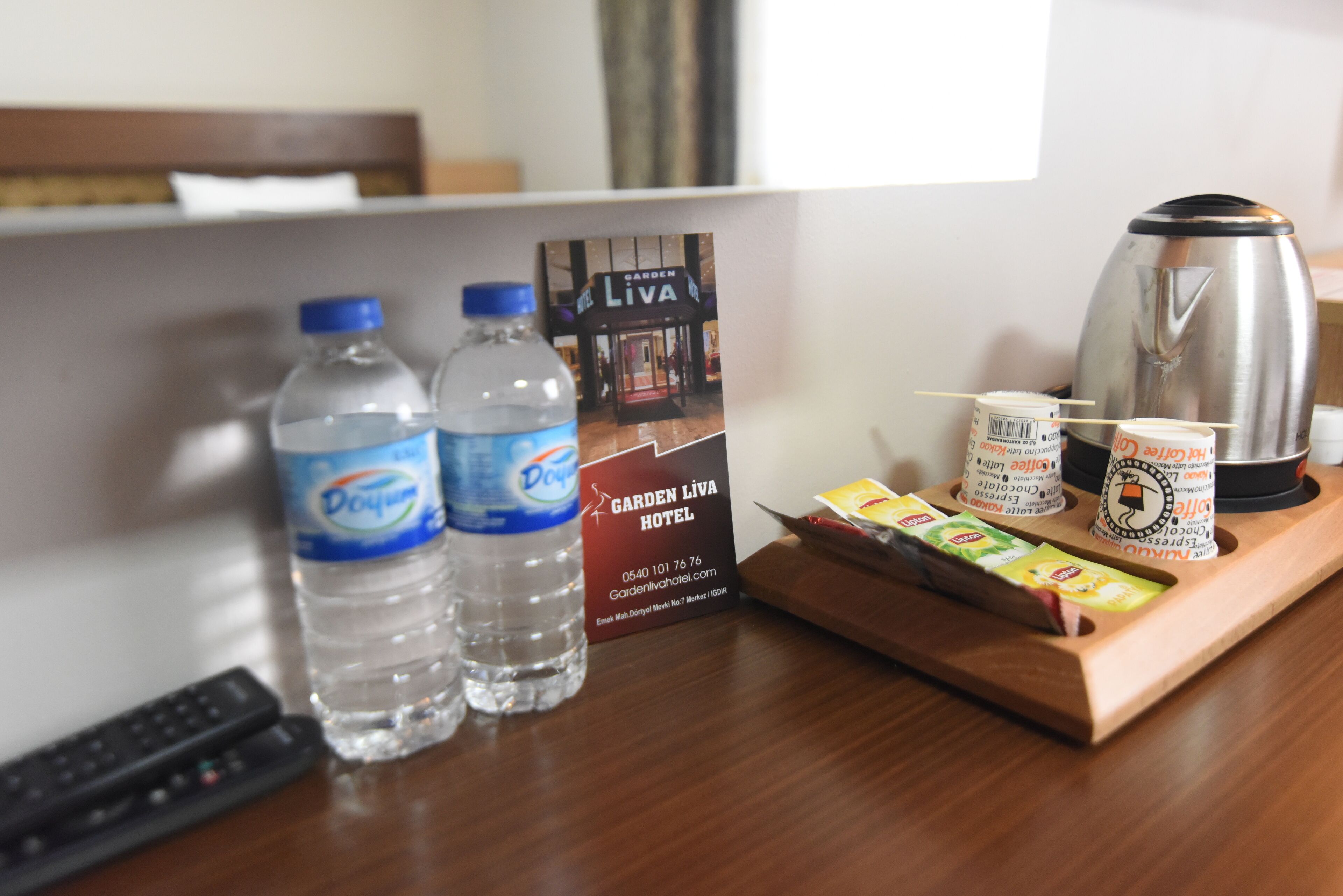 A hotel room with a remote control, two water bottles, a coffee maker, a tray with coffee packets, and a sign for Garden Liva Hotel.