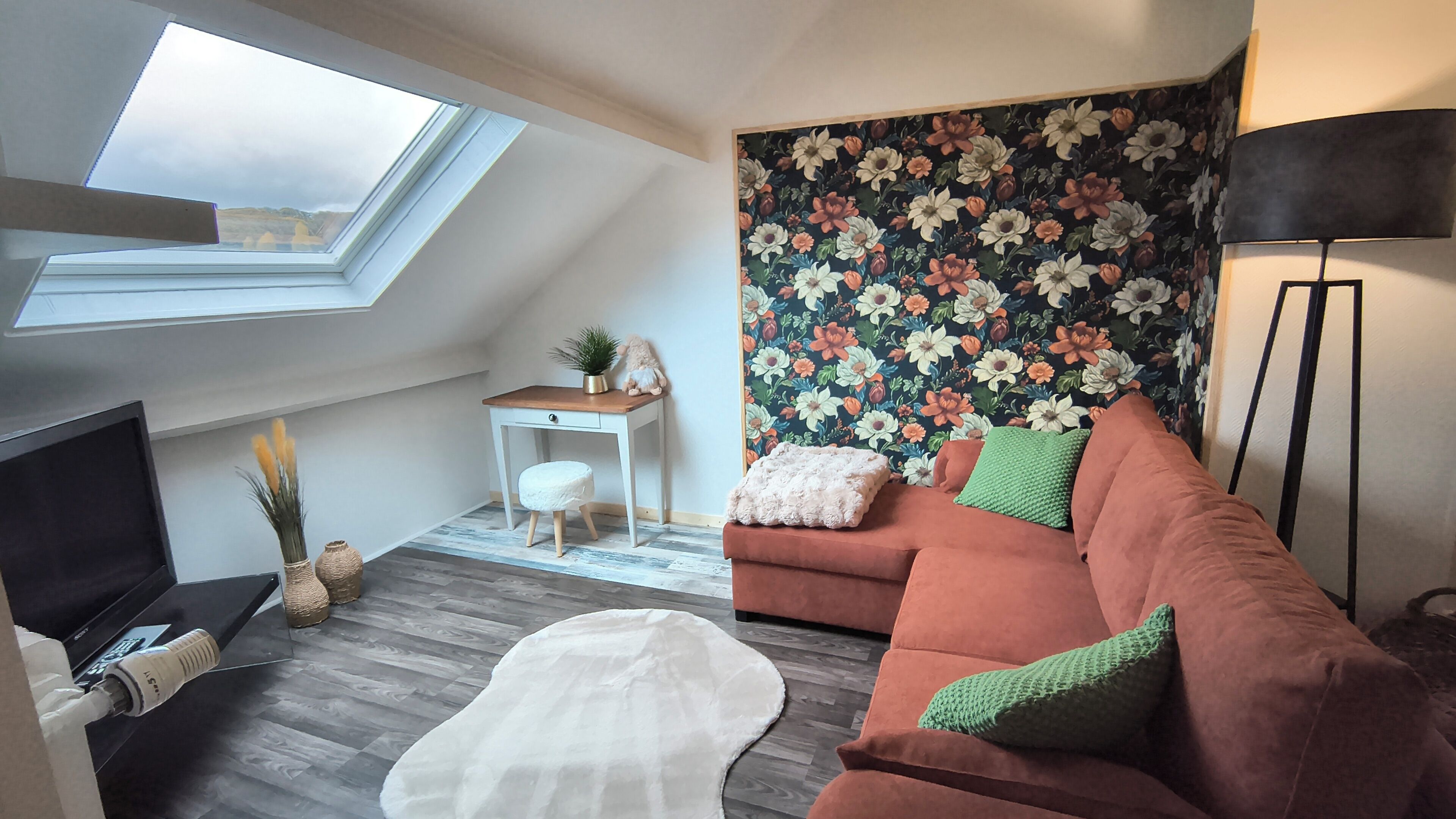 Ein Loft-Zimmer mit schrägem Dach, einer Couch, einem Fernseher, einem Schreibtisch und blumigem Wandpapier.