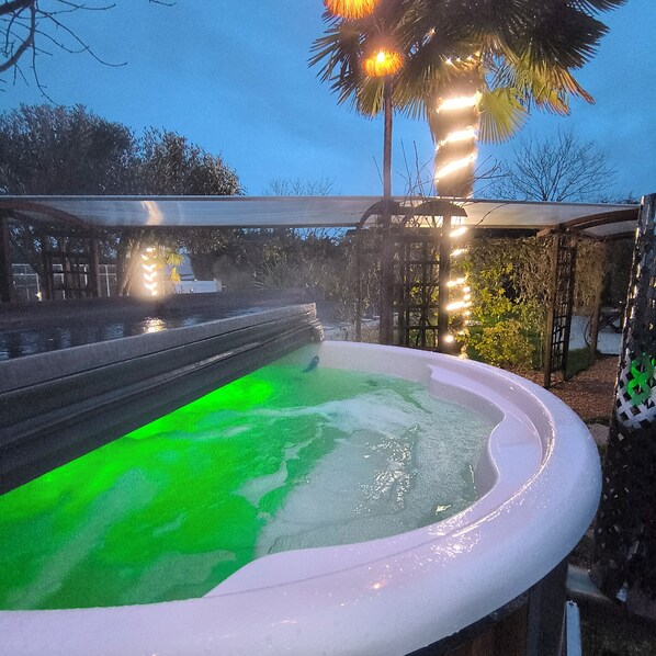 A hot tub with a green light, surrounded by trees and a covered area.