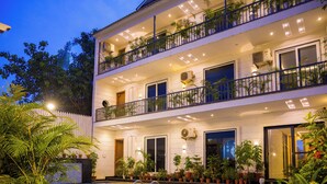 A multi-story hotel with a swimming pool, balconies with plants, and a well-lit exterior. - Alaya Stays Versace Farms |5 BHK Farm house (Noida)