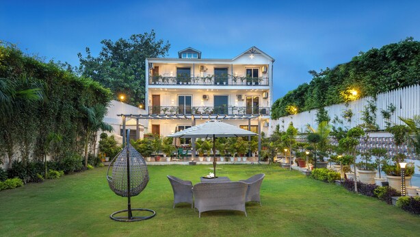 A two-story building with a balcony, surrounded by a well-maintained garden with a lawn, chairs, and a hanging chair. - Alaya Stays Versace Farms |5 BHK Farm house (Noida)