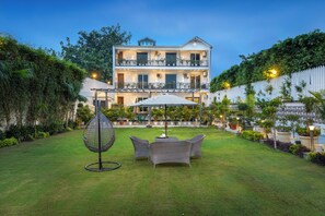 A two-story building with a balcony, surrounded by a well-maintained garden with a lawn, chairs, and a hanging chair. - Alaya Stays Versace Farms |5 BHK Farm house (Noida)