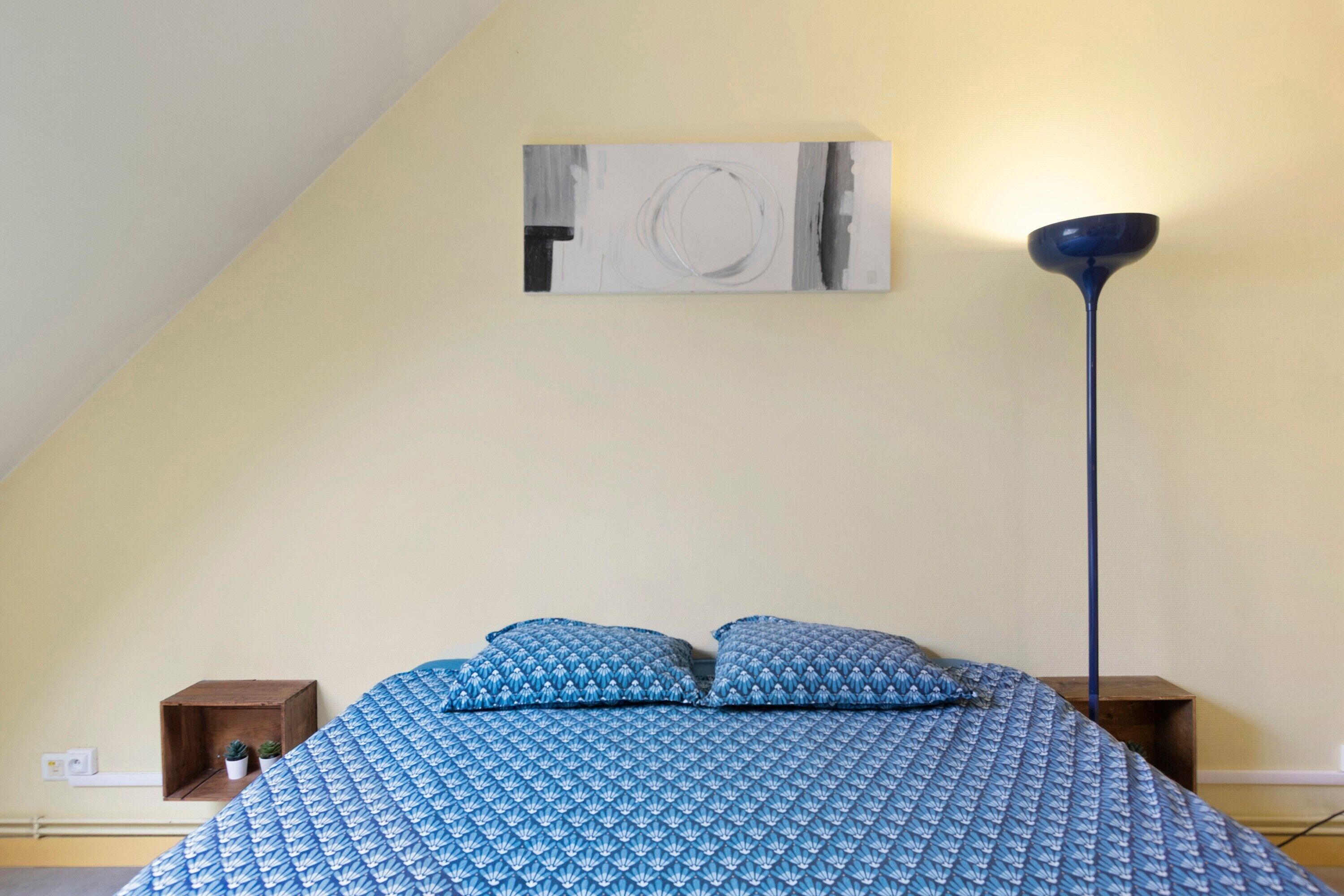 A bedroom with a bed, two pillows, a floor lamp, and a wall-mounted artwork.