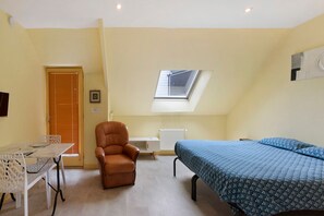 Ein kleines Zimmer mit einem Bett, einem Sessel, einem Tisch und einem Oberlicht.