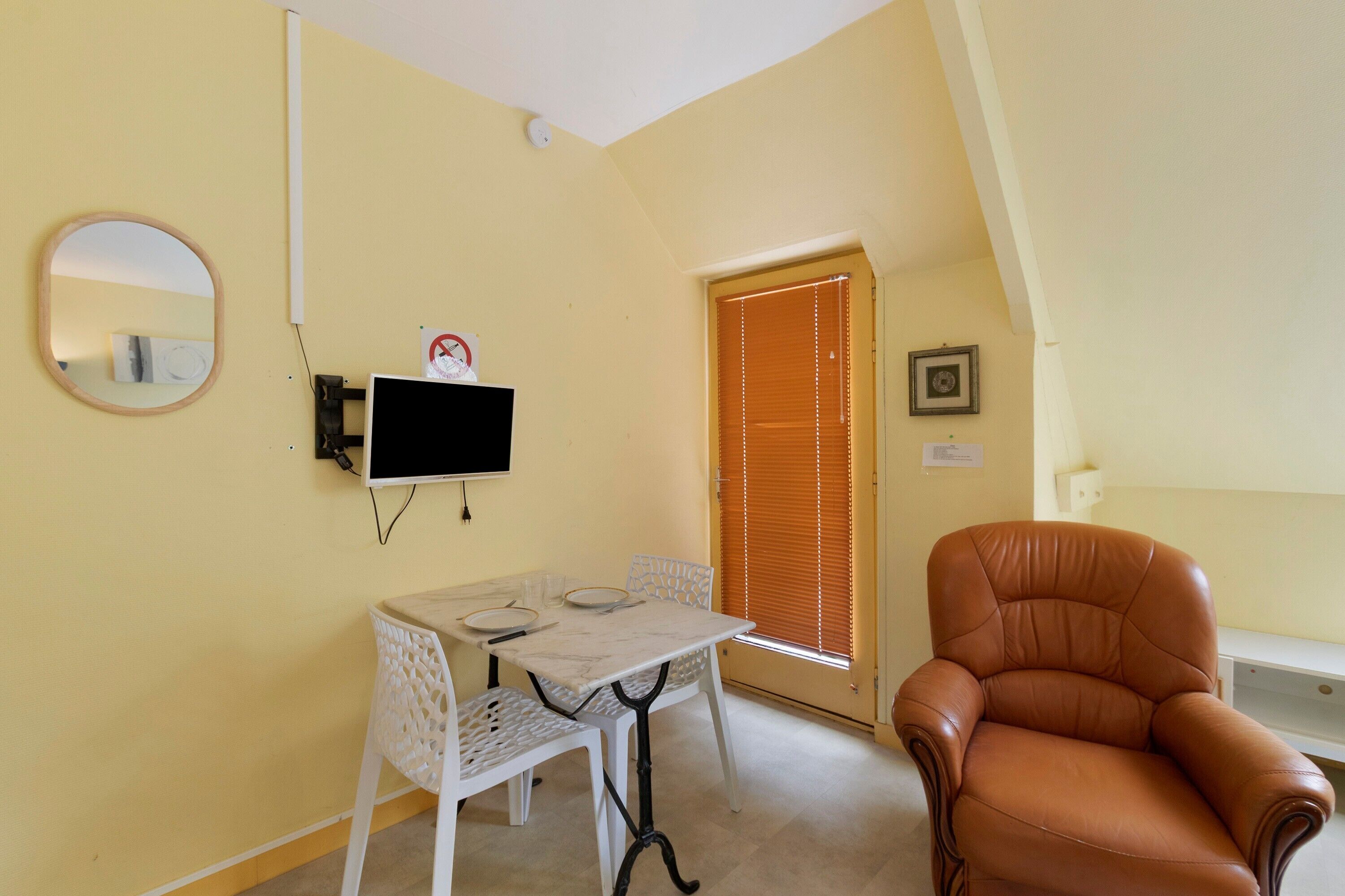 A room with a television mounted on the wall, a table with chairs, a window with blinds, and a brown leather chair.