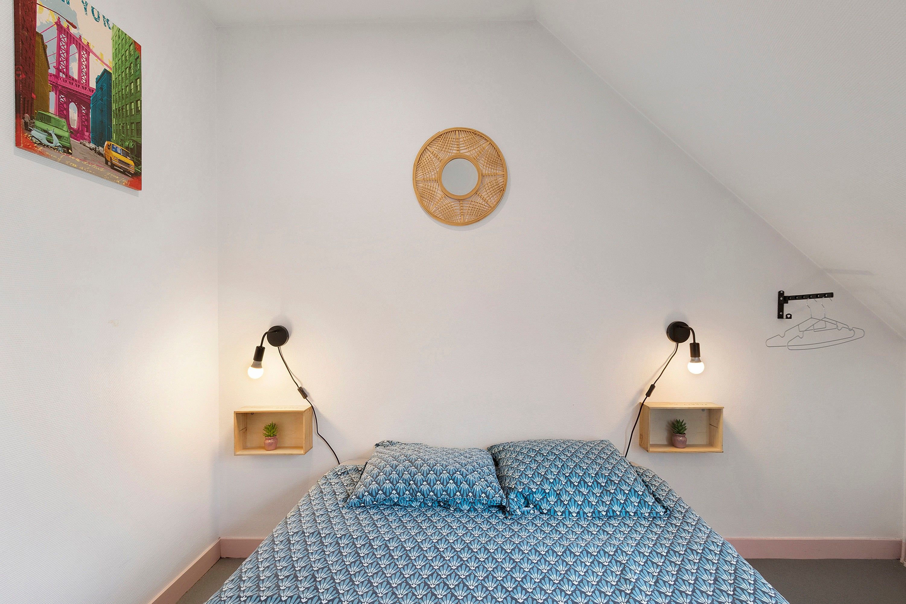 Un dormitorio con una cama, dos mesitas de noche, una lámpara de pared, un adorno circular en la pared y una planta pequeña sobre una de las mesitas.