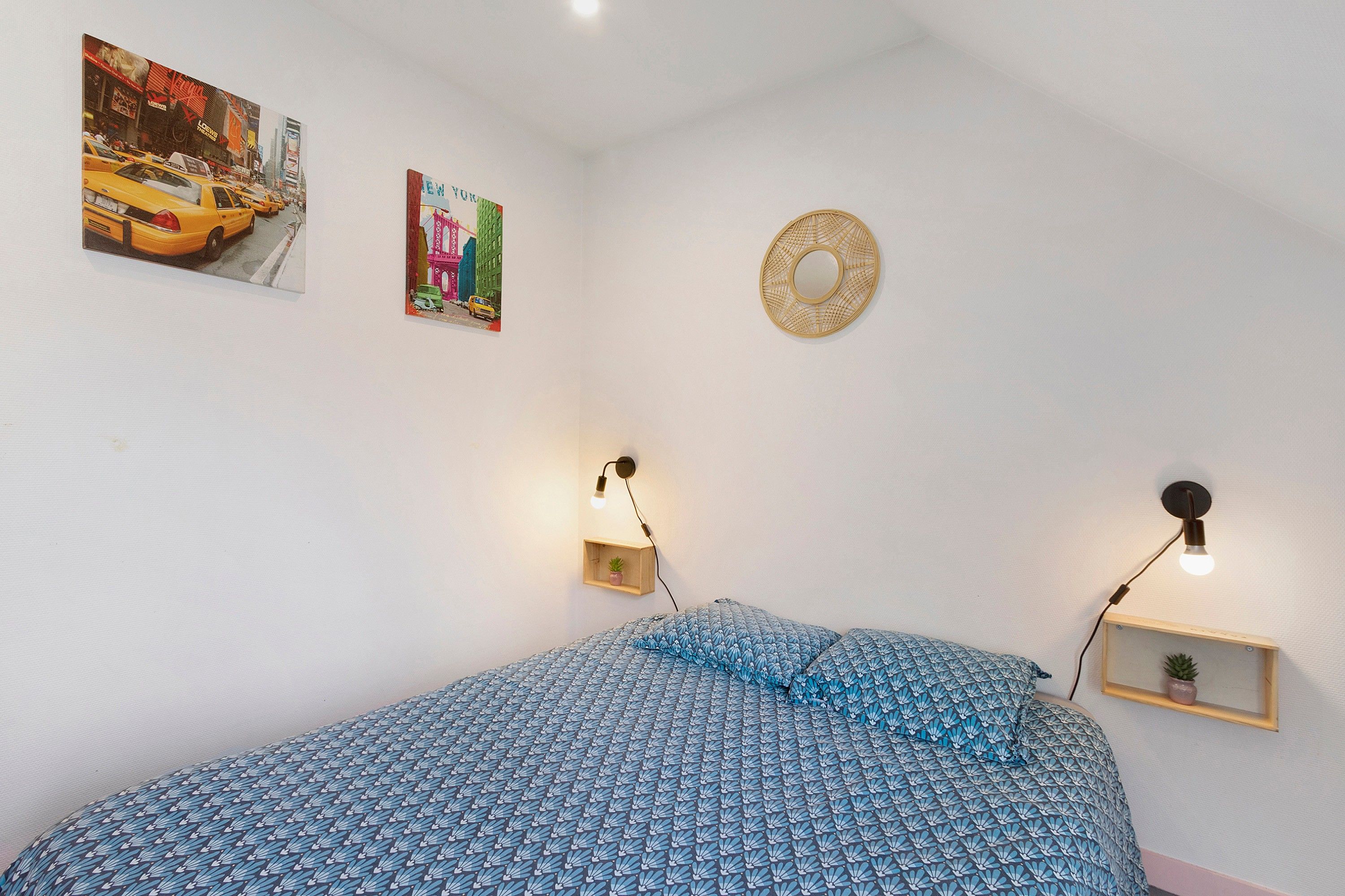 Une chambre à coucher avec un lit, deux lampes fixées au mur, une petite étagère avec une plante et deux tableaux encadrés accrochés au mur.