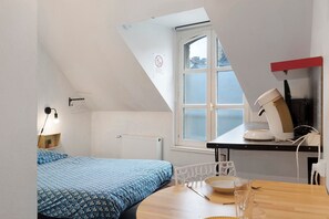 Una habitación pequeña y bien iluminada con una cama, una mesa con una silla, un radiador y una ventana con vistas a edificios.