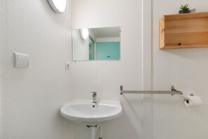 Un baño con lavamanos blanco, espejo, dispensador de papel higiénico y un pequeño estante de madera.