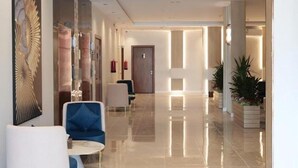 Een moderne hotelentree met een gepolijste vloer, een zithoek met een blauwe stoel en een klein tafeltje, en een groot decoratief wandkunstwerk.