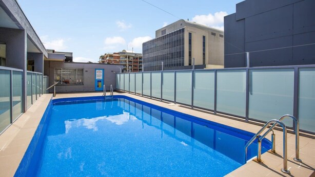 A rectangular indoor swimming pool with clear blue water, surrounded by a glass barrier and metal railings. - Chic Studio With a Pool and Moments From Shop (Sydney)