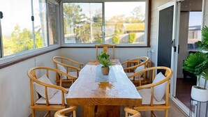 Un espace extérieur abrité, aménagé pour les repas, avec une table en bois et six chaises, une plante en pot posée sur la table, et une vue sur les arbres et les bâtiments à travers les fenêtres.