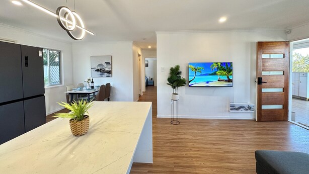 Une cuisine moderne dotée d’un grand îlot central, un coin repas avec une table dressée pour deux personnes et un espace salon avec un téléviseur et une plante.