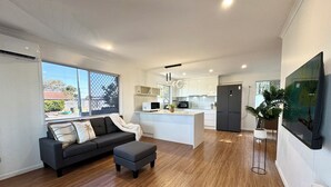 Un espace de vie moderne comprenant un canapé, un pouf, un coin cuisine et un grand téléviseur.
