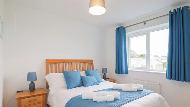 A bedroom with a wooden headboard, a bed with blue bedding, two bedside tables with lamps, a window with blue curtains, and a view of the outside. - Blissful Ocean Breeze (Perranporth)