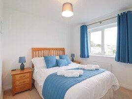 A bedroom with a wooden headboard, a bed with blue bedding, two bedside tables with lamps, a window with blue curtains, and a view of the outside.