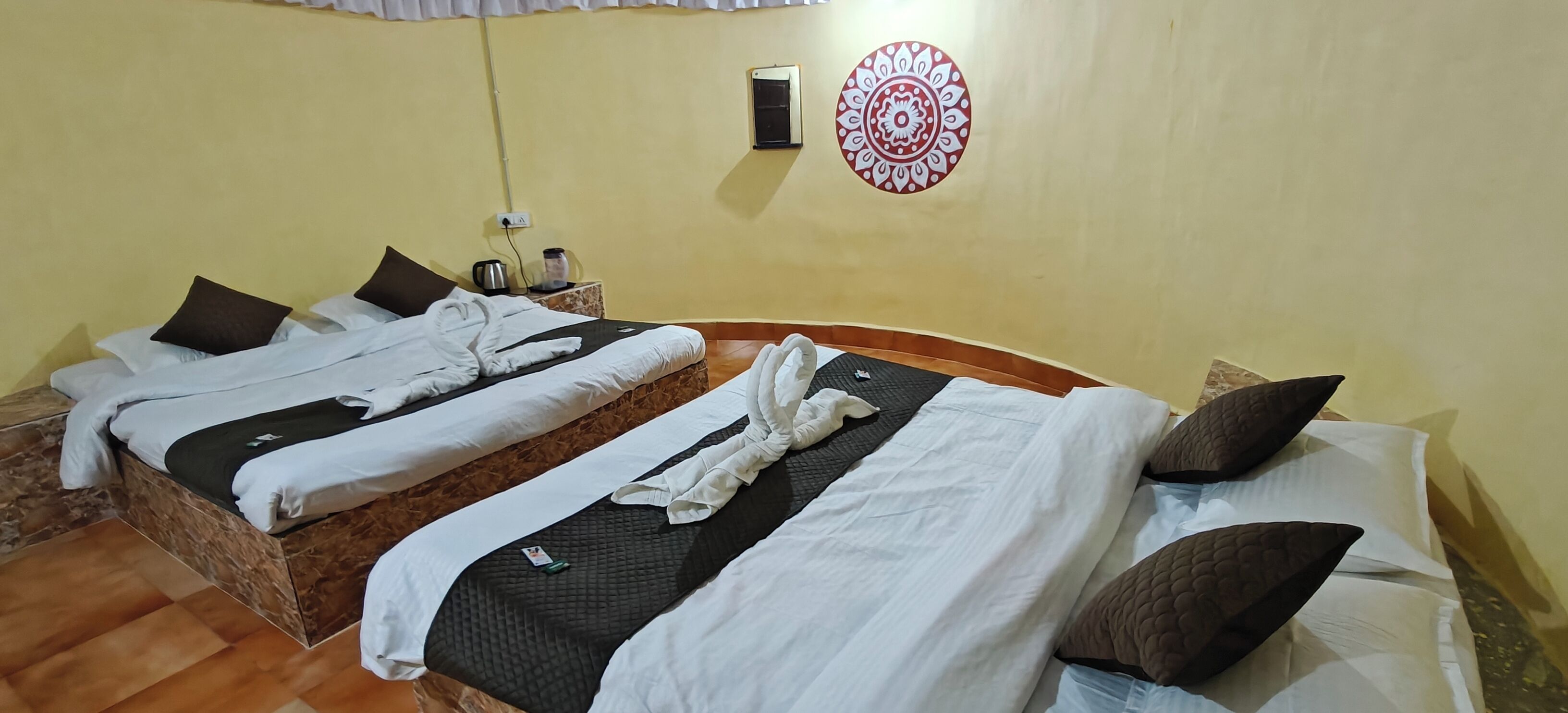 A hotel room with two beds, a wall-mounted air conditioner, a decorative wall hanging, and a small table with a kettle and a container.