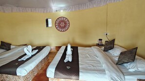 A room with two beds, each with a brown headboard, white linens, and a brown blanket. There is a wall-mounted decorative item and a small table with a lamp. - Kutch Desert Homestay (Bhuj)