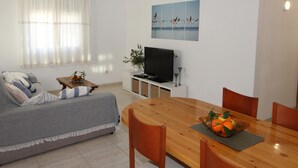 Uma sala de estar com sofá, uma mesa de jantar de madeira com cadeiras, uma TV e um quadro na parede.