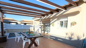 Um pátio externo com mesa e cadeiras de madeira, rodeado por uma pérgula.