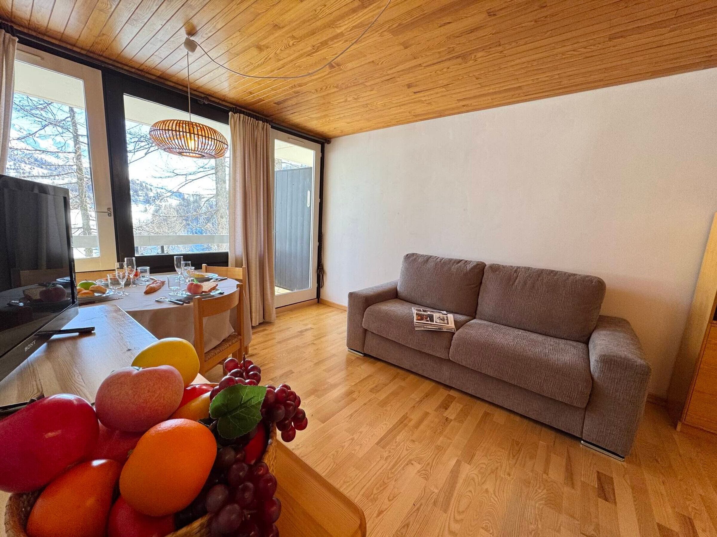 Un salon moderne avec un plafond en bois, un canapé gris, une table à manger avec des fruits et des verres, et un téléviseur.