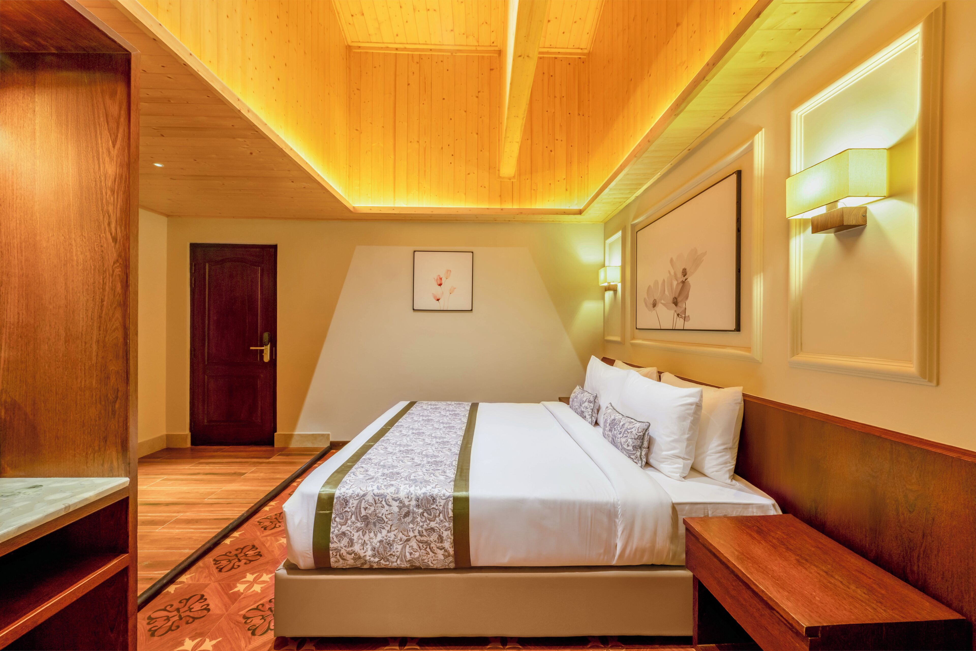 A hotel room with a wooden ceiling, a bed with white bedding, a wooden bench, a door, and a framed picture on the wall.