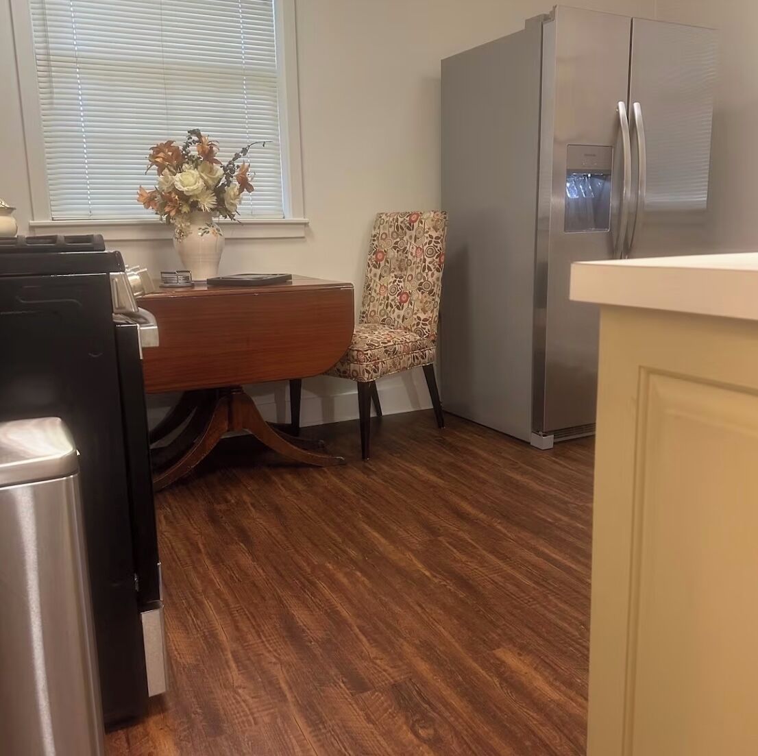 A kitchen with a wooden dining table, a patterned chair, a refrigerator, and a window with blinds.