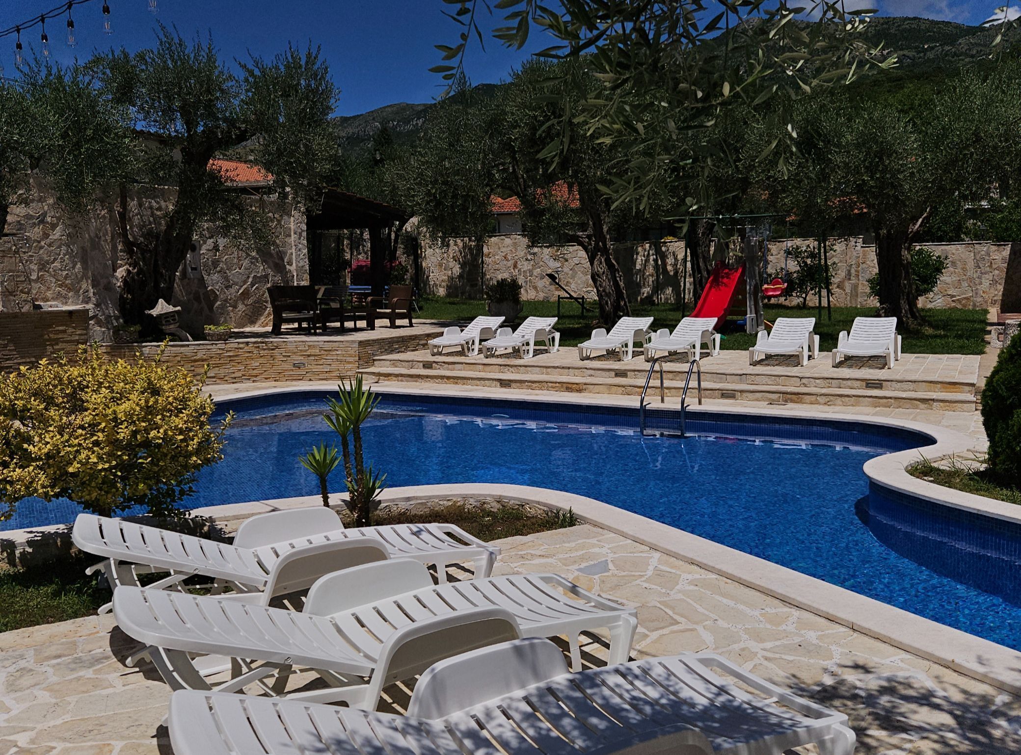 A swimming pool with lounge chairs and a slide, surrounded by stone walls and trees.