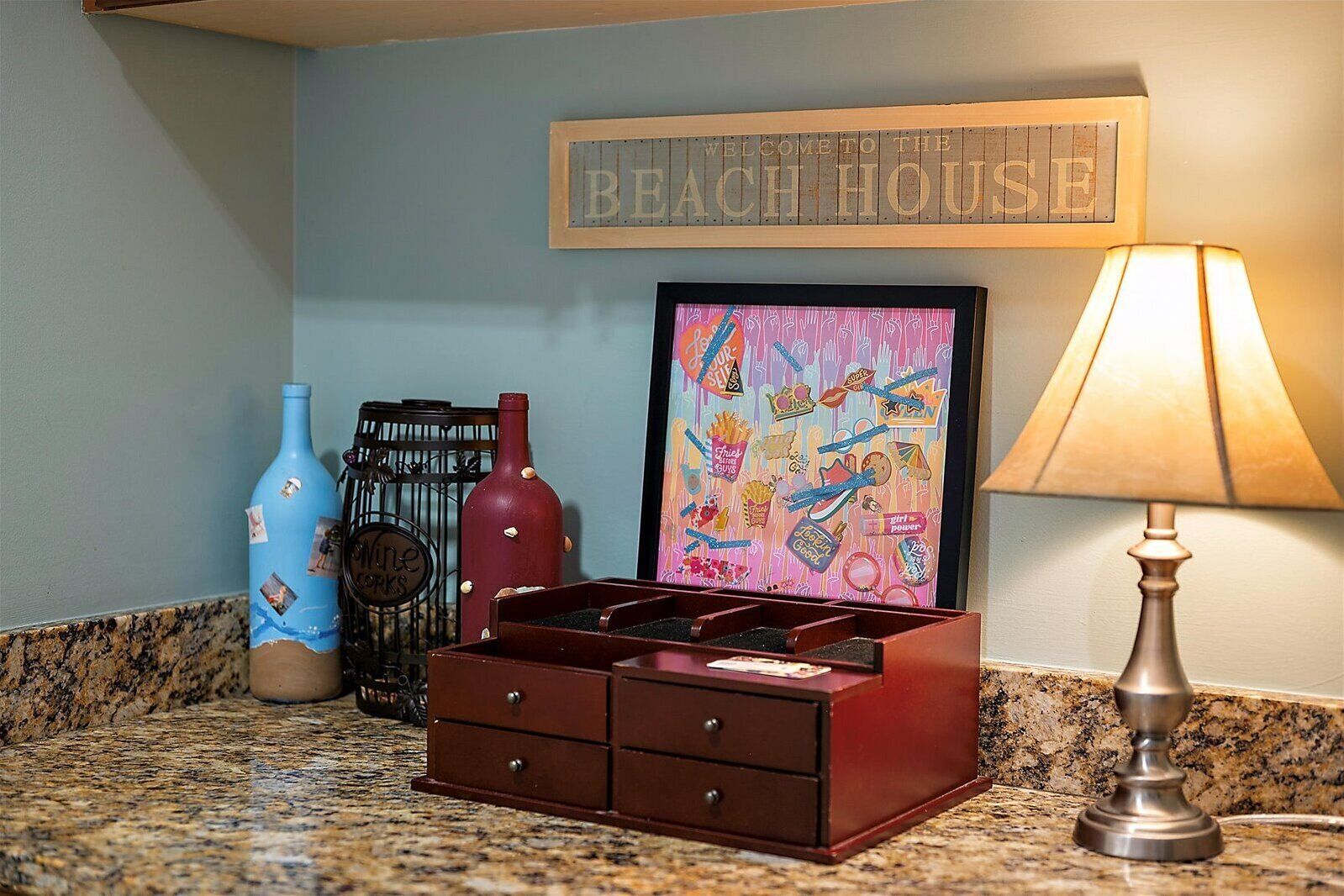 A kitchen counter with a wine rack, a framed picture, a lamp, and a sign that reads "Welcome to the Beach House".