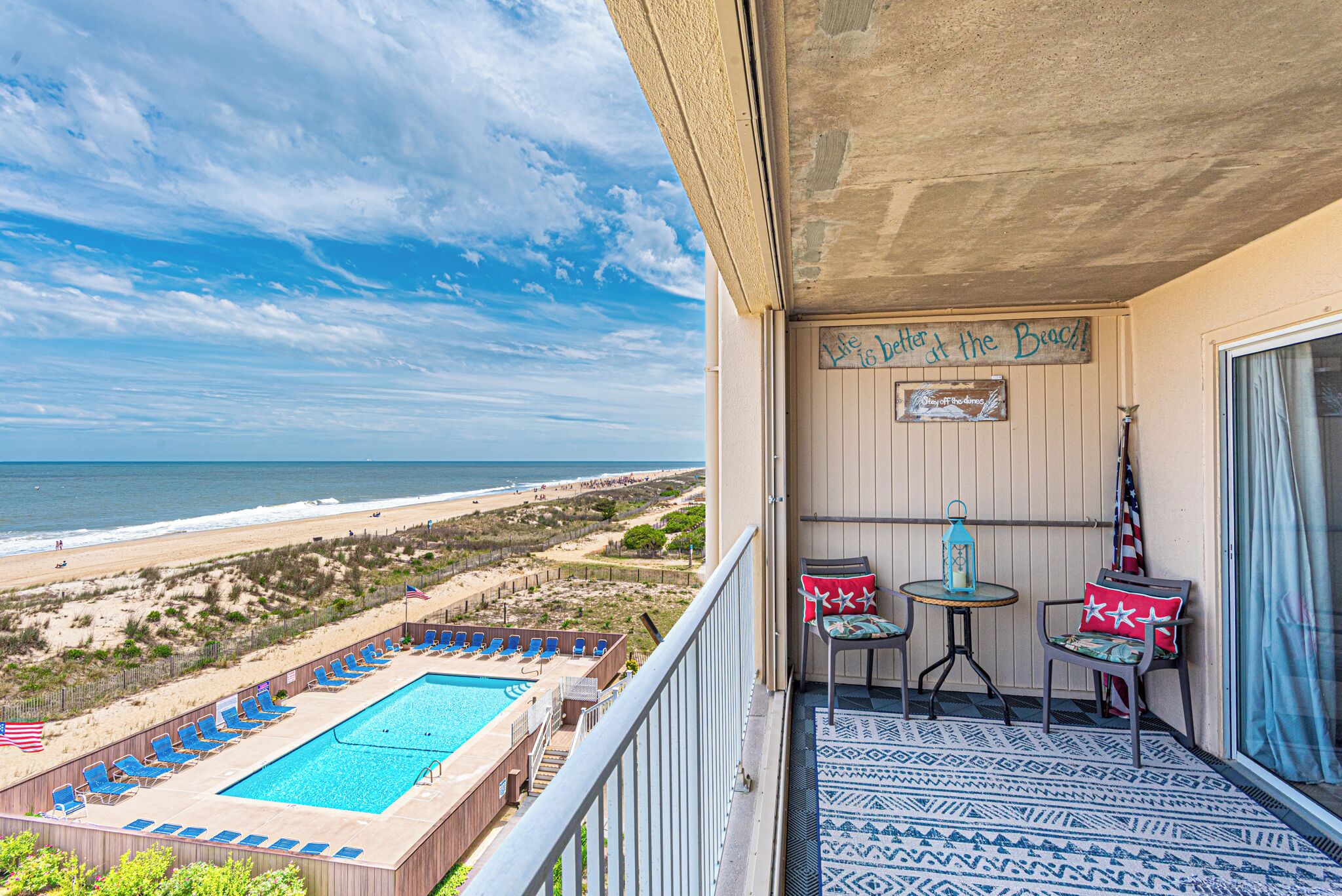 A balcony with a view of the beach, a pool, and a sign that reads 'Life is Better at the Beach.'