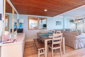 A modern living room with a wooden ceiling, a dining area with a table and chairs, a sofa, and a kitchen visible through an open door. - Beautiful remodel one Bedroom Ocean Front with outdoor pool (Ocean City)
