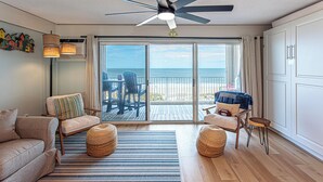Ein Zimmer mit Deckenventilator, einer Couch, einem Sessel, einem kleinen Tisch, einem gewebten Hocker und Meerblick durch große Schiebetüren.
