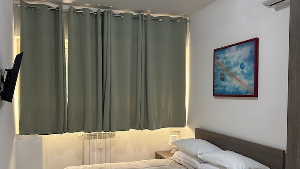 A neatly made bed with white linens, a bedside table with a lamp, a painting on the wall, and a window with curtains. - New Florence Smart Flat - Parking & Tram Stop (Firenze)
