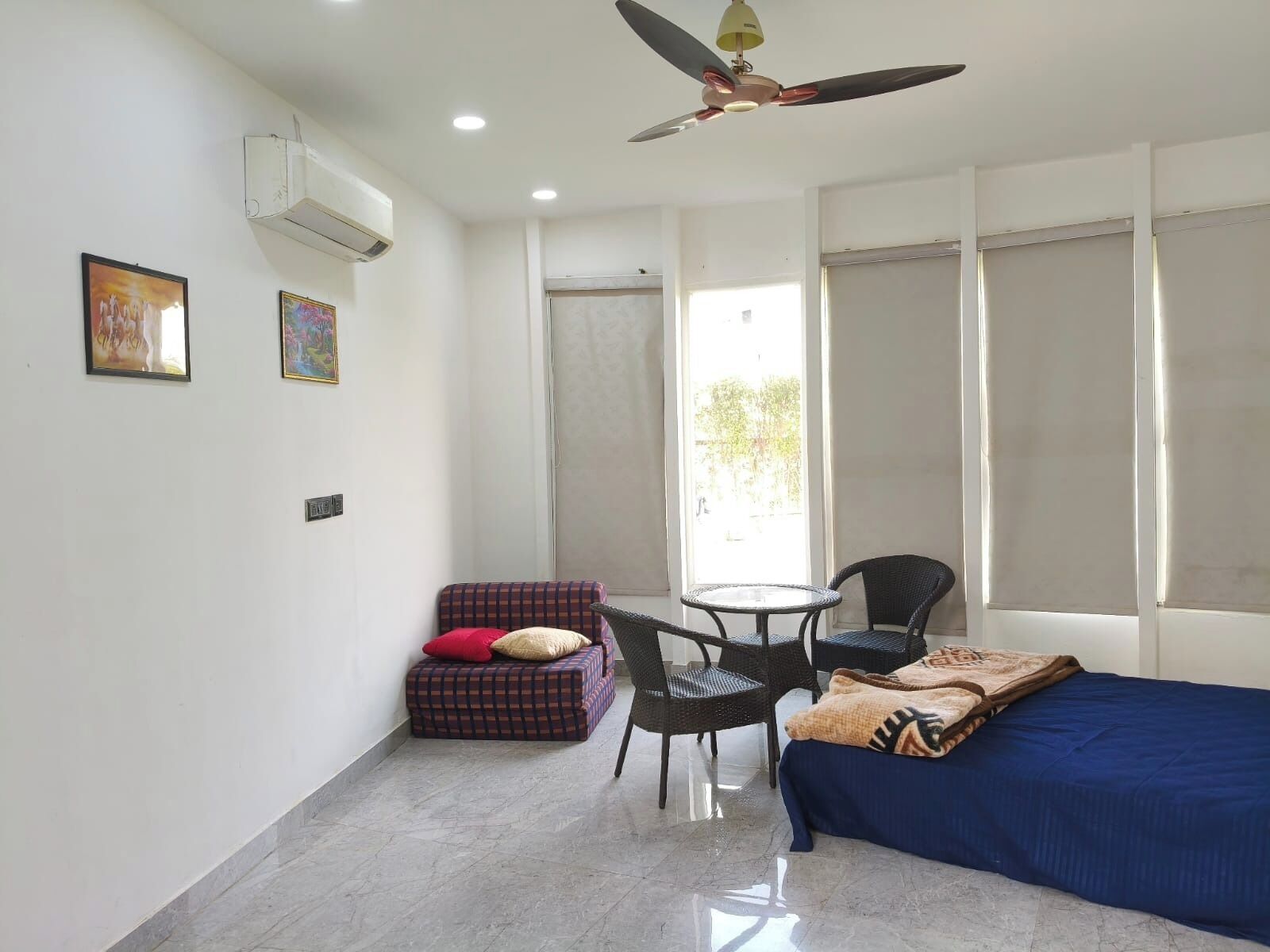 A room with a bed, a small table, two chairs, a ceiling fan, and a window with blinds.