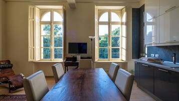 A dining area with a long wooden table, chairs, a television, and windows with a view of trees.
