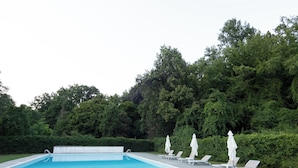 Uma piscina retangular com superfície azul translúcida, ladeada por um deck pavimentado. Há várias espreguiçadeiras brancas com guarda-sóis, e a área é cercada por árvores e arbustos verdes exuberantes.