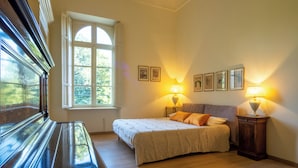 Quarto com cama, duas luminárias de cabeceira, mesa de cabeceira de madeira, janela com cortinas e um piano.