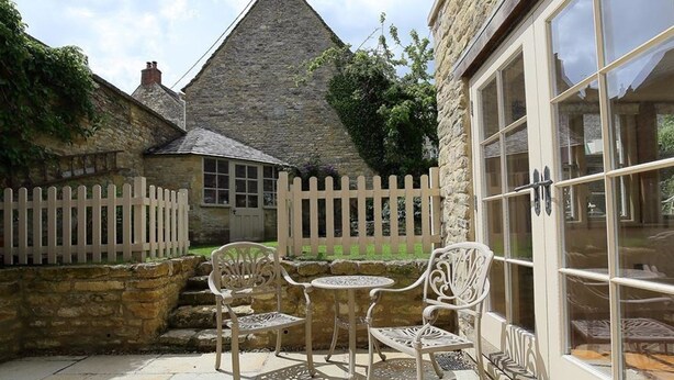 A stone building with a triangular roof, a white picket fence, and a patio with two chairs and a table. - The Coach House (Burford)