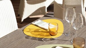Un coin repas avec des chaises en osier, une table dressée avec des sets de table jaunes, des verres à vin et un menu.
