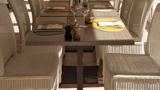 Un espace repas avec une longue table dressée pour six personnes, entourée de chaises en osier, et offrant une vue sur l’extérieur grâce à de grandes fenêtres.