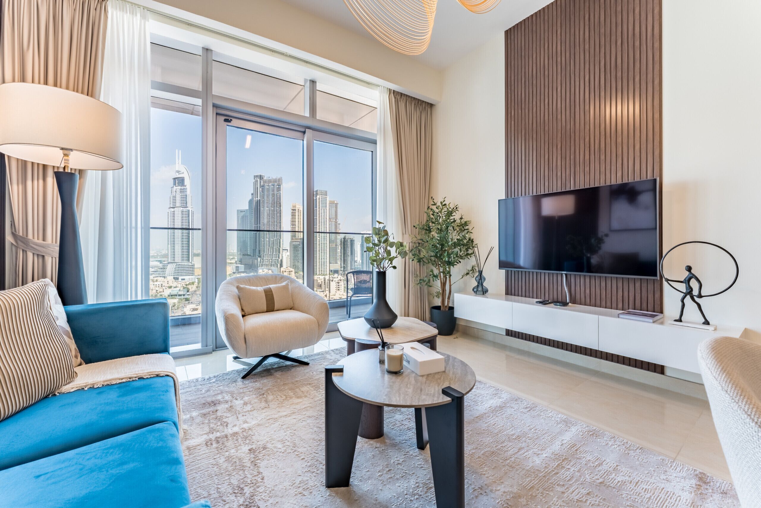 A modern living room with a large TV, a blue sofa, a beige armchair, and a view of the city skyline through floor-to-ceiling windows.