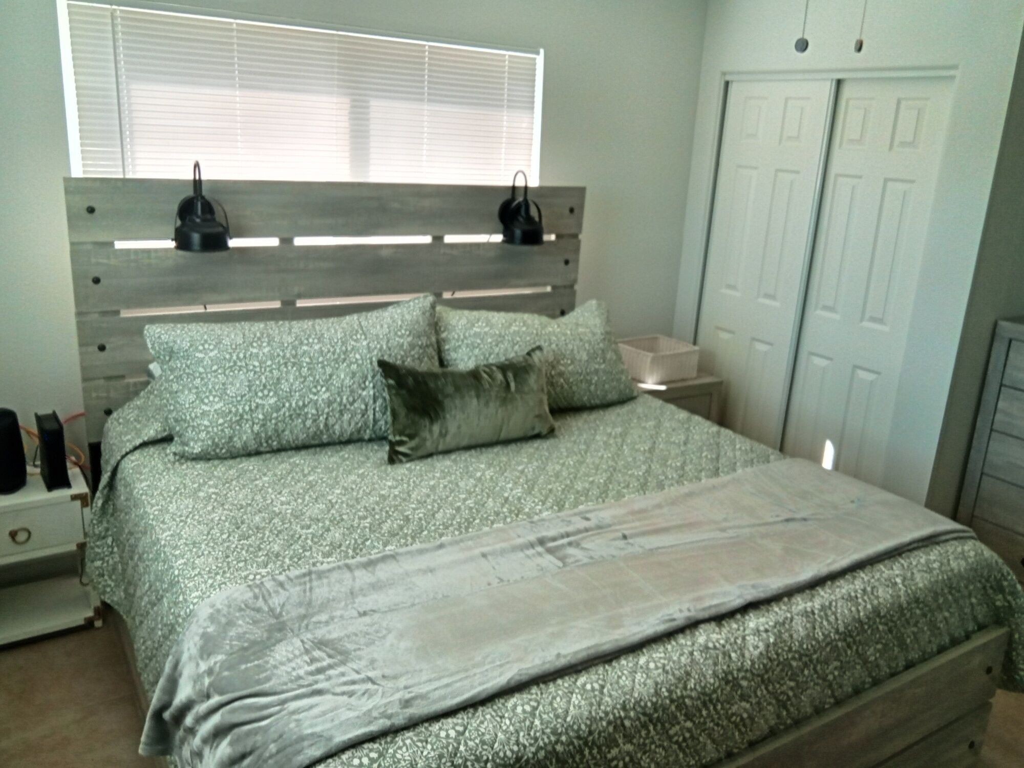 A bedroom with a bed, a nightstand, a dresser, a window with blinds, and a door.