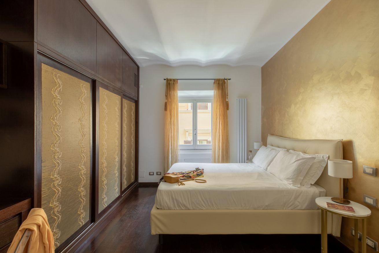 A hotel room with a large bed, a wardrobe, a window with curtains, and a bedside table with a lamp.
