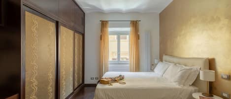 A hotel room with a large bed, a wardrobe, a window with curtains, and a bedside table with a lamp.