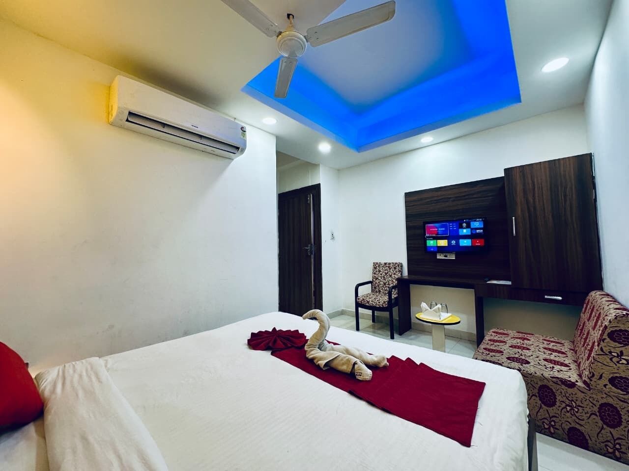 A hotel room with a bed, a ceiling fan, an air conditioner, a TV, a small table, and a sofa.
