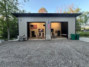 A building with two open doors, an American flag, a grill outside, and two green trash bins. - Sleeps 14, Hot Tub, Garage Event Room, Game Room Lake + River + Trails + Golf (West Branch)